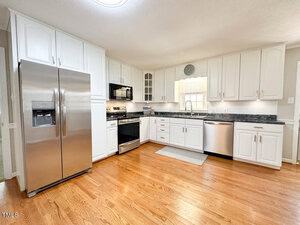 A modern kitchen with white cabinets, stainless steel appliances, and a wooden floor. It features a refrigerator, oven, microwave, dishwasher, and a window above the sink. The countertops are dark, providing a contrast to the light cabinetry.