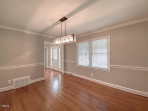 A spacious dining room with light gray walls, a modern rectangular light fixture, and hardwood floors. A large window with blinds allows natural light to enter. A doorway leads to another room, and there's a white door in the background.