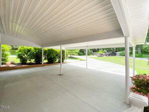 Covered patio with white ceiling and floor, supported by poles. Green shrubs on the left and a driveway leading to a parked car in the background. Lush greenery and a clear sky are visible beyond the patio.
