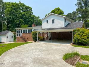 A suburban two-story house with white siding and brick walls, featuring an attached carport. The front yard has a well-maintained lawn, a small shed, and is surrounded by lush greenery and trees. The driveway is wide and paved.