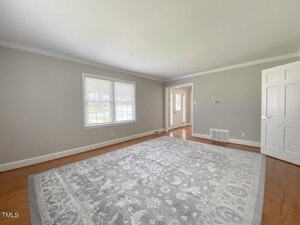 A spacious, empty room with light gray walls and a large window allowing natural light. The hardwood floor features a large, ornate gray and white rug. An open doorway leads to another room.