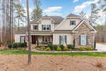 A large, modern two-story house at 3597 Carole Court, Wake Forest, boasts stone and beige siding surrounded by a landscaped yard. An American flag proudly flutters near the front porch adorned with red chairs. Tall trees and a partly cloudy sky serve as the serene backdrop.