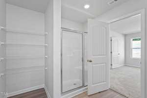 A minimalist bathroom with white walls features a glass-enclosed shower next to open wire shelving. The door is open to a bright carpeted bedroom with two windows, allowing natural light in, illuminating the wooden floor.