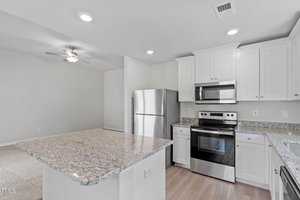 Modern kitchen with white cabinets, stainless steel appliances including a fridge, oven, and microwave. Features granite countertops and a center island. Adjacent open living area with a ceiling fan and light wood flooring.