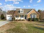 The single-story house at 3527 Bluebonnet Drive in Wake Forest boasts a charming stone and siding exterior with a gabled roof. A covered front porch with white railings is framed by shrubs, and the driveway leads to an attached garage. The sky above is partly cloudy, completing this picturesque setting.