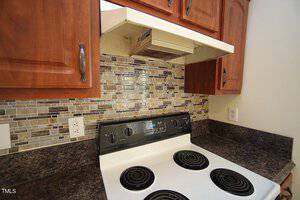 A kitchen with wooden cabinets, a mosaic tile backsplash, and a white electric stove with black burners. The countertop is dark, and there's a range hood above the stove. Two electrical outlets are visible on the backsplash.