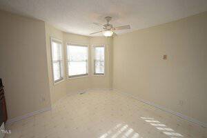A bright, empty room with light-colored walls and a white ceiling fan. Three large windows with blinds allow natural light to enter, illuminating the tiled floor.