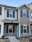 A two-story townhouse at 102 Winston Way in Creedmoor boasts gray and beige siding, black shutters, and a bright blue front door. It features two sets of windows on each floor and a small front yard with a concrete walkway. A real estate sign is prominently displayed on the lawn.