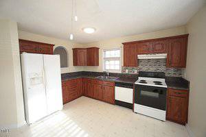 A spacious kitchen with dark wooden cabinets, black granite countertops, and a tiled backsplash. It features a white refrigerator, a white oven with a black hood, and a dishwasher. The floor is light-colored tile, and there is a window above the sink.