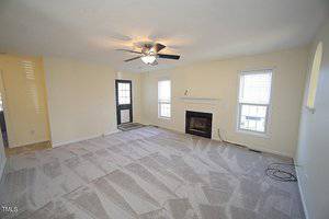 A spacious living room with beige carpet and cream walls. There is a ceiling fan with lights, a fireplace with a white mantle, and two windows with blinds. A green door with glass panels leads outside, and an arched opening is visible on the right.