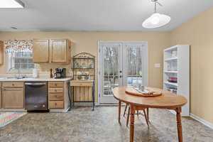 A cozy kitchen features wooden cabinets, a countertop with a sink, and a coffee maker. A round wooden table has a plate of food. French doors lead outside, next to a white bookshelf and wall-mounted light. A colorful rug adds a touch of brightness.