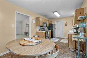 A cozy kitchen with wooden cabinets and a round dining table. The room includes a black refrigerator, stove, and various kitchen essentials on the countertops. Colorful rugs are on the floor, and an open door leads to another room.