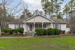 Nestled on 1410 W B Street, this single-story house in Butner showcases a charming front porch surrounded by trimmed bushes and a lush lawn. An American flag proudly hangs by the entrance, with tall trees and a partly cloudy sky as its serene backdrop.