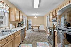 A cozy kitchen with wooden cabinets, a double sink, and black appliances. A small dining area with a table and chairs sits near a window, with a ceiling light above. Light walls and floral curtains add warmth to the space.