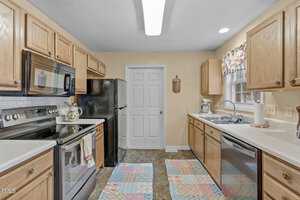 A cozy kitchen with wooden cabinets, stainless steel appliances, and a patchwork-style patterned rug on a tiled floor. The room features a window with floral curtains, a microwave, and a stand mixer on the counter.