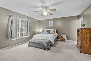 A bedroom with olive green walls features a bed with white and gray bedding, two nightstands with lamps, a wooden dresser, and a ceiling fan. A window with gray curtains allows natural light in. A small table with a lace cover is in the corner.