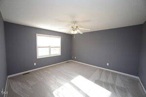 An empty room with gray walls and a light-colored carpet. A ceiling fan with lights is centered in the room. The room has a large window on one wall, allowing natural light to enter.