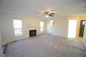 A spacious living room with beige carpet, a ceiling fan, and a fireplace. Two windows bring in natural light. There's an open doorway on the right leading to a sunlit room. Walls are painted in a light cream color.