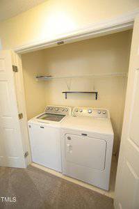 A small laundry nook with a white washing machine and dryer side by side. Above them is a wire shelf and a small black shelf for storage. The area is enclosed by open white folding doors with beige walls surrounding the appliances.