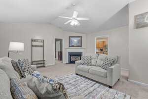 A spacious living room with a vaulted ceiling and a ceiling fan. There's a fireplace with a framed picture above it, surrounded by two beige sofas with patterned cushions. A lamp and a patterned rug add to the cozy ambiance. An open doorway leads to a kitchen.