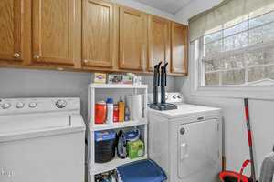 A laundry room with a washing machine and dryer, wooden cabinets, a small white shelf holding detergent and cleaning supplies, and a window with a green valance. A vacuum cleaner and a broom are nearby.