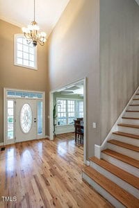 Bright foyer with high ceilings and chandelier, featuring a wooden staircase on the right, an oval glass front door, and a view into a dining area with a table and chairs. Sunlight filters through large windows, illuminating the hardwood floor.