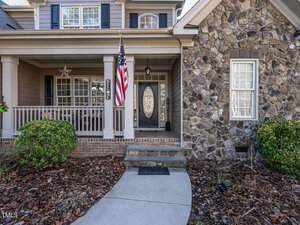 A charming house with a stone facade and a front porch featuring a decorative railing. An American flag hangs beside the entrance, which has a door with an oval glass window. The pathway is bordered by mulch and shrubs.