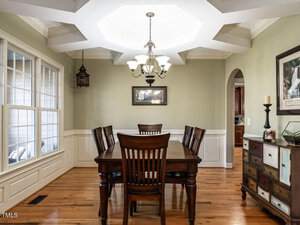 Dining room with a wooden table and six chairs. A chandelier hangs from the coffered ceiling. The walls are painted green with white wainscoting. Large windows provide natural light, and there's a sideboard with decorations on the right.