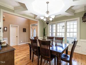 A dining room with a dark wooden table and six chairs, under a decorative chandelier. The room has large windows with white trim, hardwood floors, and a plant in the corner. The walls are light-colored with wainscoting design.
