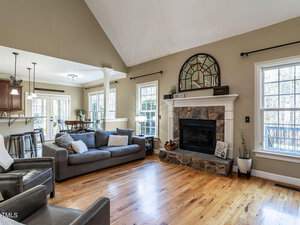 A cozy living room with wooden floors and a large stone fireplace topped with a decorative mirror. A gray sofa, armchair, and a side table furnish the space. Natural light streams in from tall windows and glass doors leading to an outdoor area.