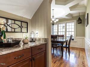 A kitchen features a granite countertop with a tray holding a wine bottle and glasses. A decorative mirror hangs above. In the background, a dining area with a wooden table and chairs is visible, lit by a chandelier and large windows.
