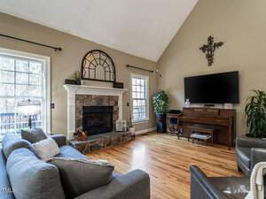 A cozy living room with a stone fireplace and a mirror above it. A large TV is mounted on the adjacent wall above a wooden stand. There are two large windows, a gray couch, armchairs, and plants decorating the space. A cross decorates the wall.