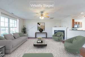 A virtually staged living room with a gray sofa, green armchairs, and a coffee table on a patterned rug. A ceiling fan is above, and a fireplace is on the right. Large windows allow natural light in, highlighting the modern decor.