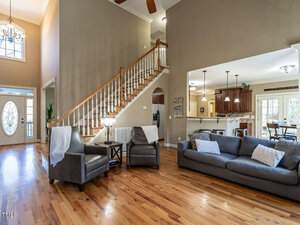 Spacious living room with a high ceiling, featuring a gray sofa, two armchairs, and a wooden coffee table. A staircase with white railings leads upstairs. Open kitchen area visible with dark wood cabinets and modern light fixtures.