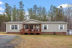Located at 6626 Woodlawn Drive in Oxford, this charming single-story house features light gray siding and a welcoming front porch with wooden steps. Nestled amid tall pine trees, it boasts a grassy yard and gravel driveway. A bicycle leans casually against the house under a partly cloudy sky.