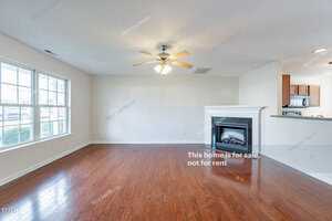 A bright living room with hardwood floors, a ceiling fan, and a fireplace. Large windows let in natural light. The kitchen is visible in the background. Text on the image reads, "This home is for sale, not for rent.