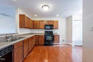 A modern kitchen with wooden cabinets, black appliances including an oven and dishwasher, and a double sink. The space features hardwood floors and bright lighting. Walls have "Not for Rent" notices.