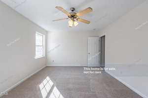 Bright, empty room with beige carpet, cream walls, and a ceiling fan with lights. A large window lets in natural light, and a white door is visible on the right. Text overlay indicates the home is not for rent.