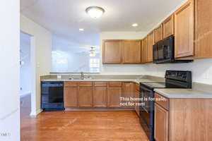 Spacious kitchen with wooden cabinets, black appliances, and hardwood floors. Walls have "Not for sale, not for rent" text. An open doorway leads to a living area with a ceiling fan and a white door.