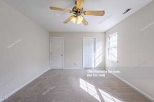 An empty carpeted room with beige walls, a ceiling fan with lights, two closed white doors, and a window letting in natural light. The wall reads, "This home is for sale, not for rent.