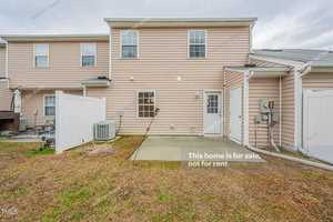 Rear view of a two-story beige house with white trim and a small concrete patio. The yard is grass-covered, and a white fence separates the property from neighbors. A "For Sale" sign is visible. Overcast sky in the background.