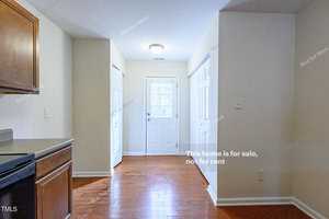 A bright, empty room with wooden floors and white walls. There are two white doors, one with a window, with "This home is for sale, not for rent" written on the image. A partial view of a kitchen counter is visible on the left.