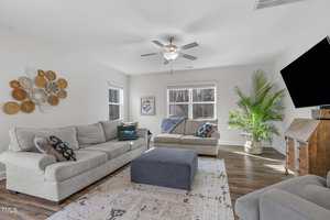 A modern living room with light gray sofas, a blue ottoman, and a patterned rug. Wall decor includes a circular art piece. A ceiling fan is above, and a flat-screen TV is mounted on the wall. Large windows and a potted plant add brightness to the space.
