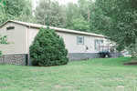 A light green mobile home on 1626 Fawn Lane boasts a wooden deck and several windows, embraced by lush greenery and trees. The stone-lined foundation complements the small satellite dish, while a well-kept lawn graces the foreground in this serene Creedmoor setting.