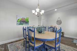 A dining room with a round wooden table surrounded by blue chairs. A chandelier hangs above the table, which holds a potted plant. A painting is on the wall, and a circular mirror is mounted nearby. A rug is on the wooden floor.