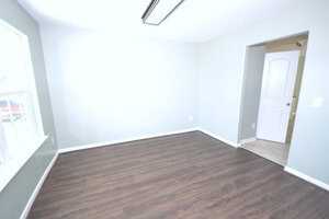 An empty room with light gray walls and dark wood flooring. A large window is on the left, and an open doorway with a white door leads to another room. A fluorescent light fixture is on the ceiling.