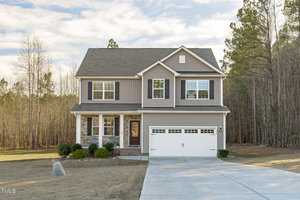 A two-story suburban house at 3304 Wiltshire Way in Creedmoor, featuring beige siding and stone accents. It includes a double garage, a vibrant red front door, and a well-maintained driveway. Nestled among trees and set against a clear sky, this home radiates charm.