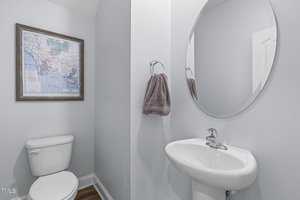 A small bathroom with light blue walls features a round mirror above a white pedestal sink and a towel hanging on a ring. A framed map artwork hangs above the white toilet, with hardwood flooring completing the decor.