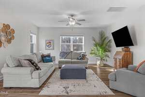 A cozy living room with two light gray sofas, an armchair, and a dark blue ottoman on a patterned rug. A ceiling fan is overhead, and a TV is mounted on the wall. The room is decorated with woven wall art and a large potted plant by the window.