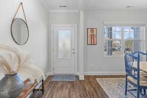 A cozy entryway with a round mirror, pampas grass in a vase on a wooden bench, a textured glass door, and an adjacent dining area with blue chairs by a window. Artwork hangs on a light wall.